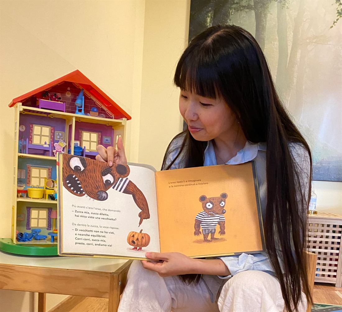 Valentina legge un albo illustrato a voce alta nel suo studio, con una casa delle bambole alle spalle.